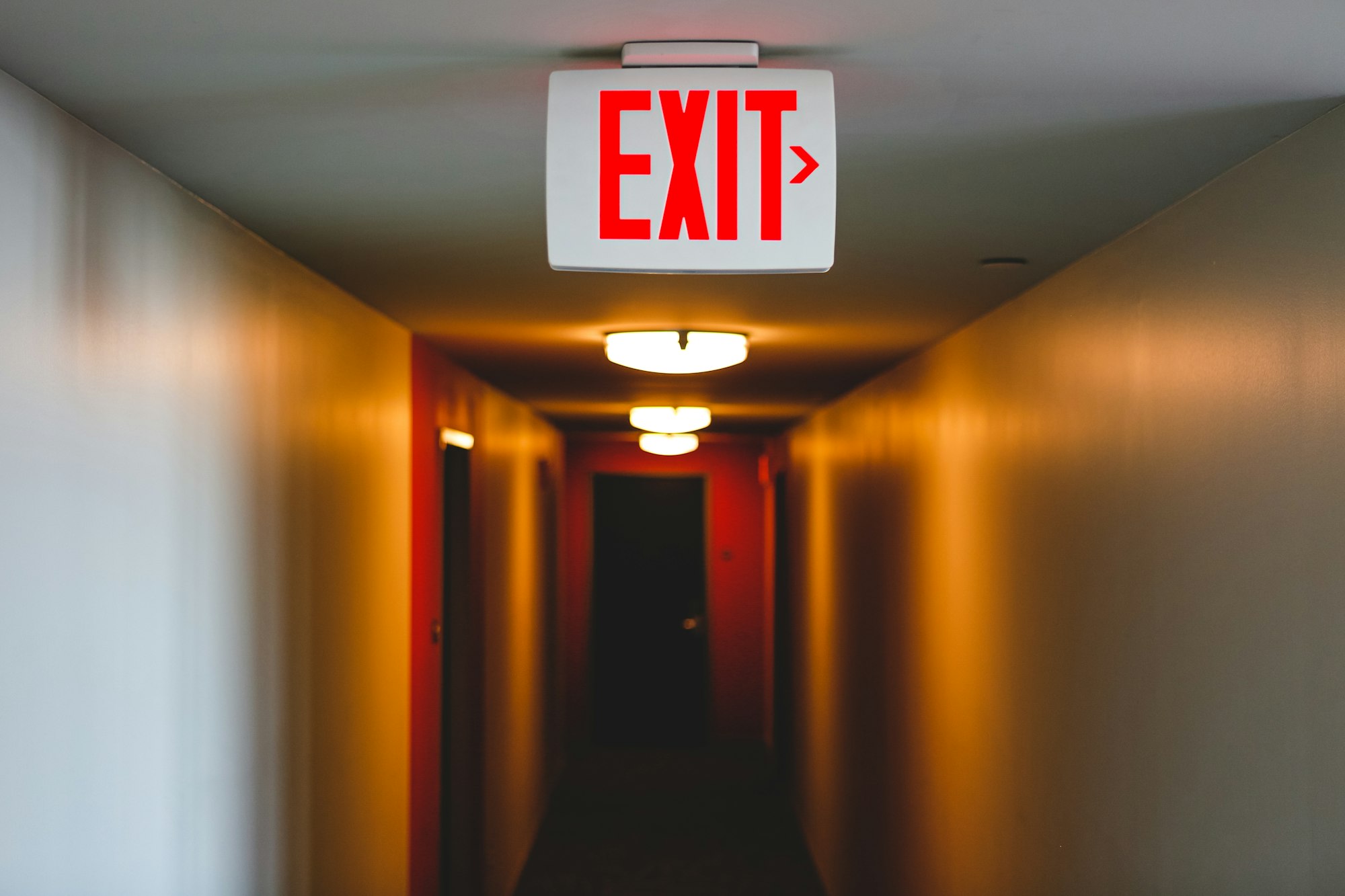 Exit sign in a lit up long hallway, exit to right from eerie hallway